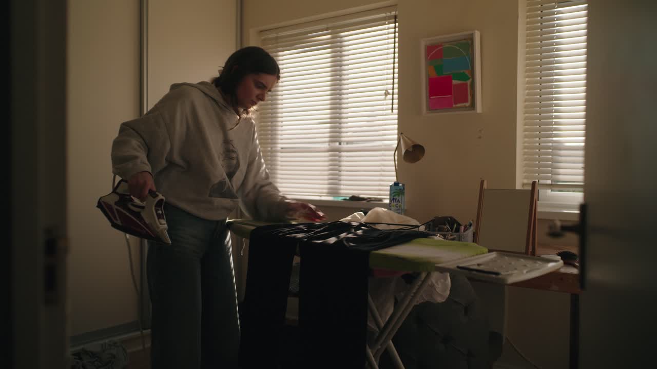 Cinematic shot of a woman ironing in a softly lit bedroom. Natural morning light through blinds adds warmth and realism to this domestic Irish interior.