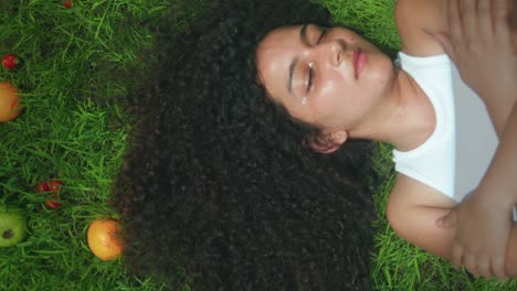 top-down drone shot of woman with voluminous curly hair and brown skin lying in green grass beside scattered fruit, eyes closed and arms crossed gently, convey serenity, connection, earthbound rest