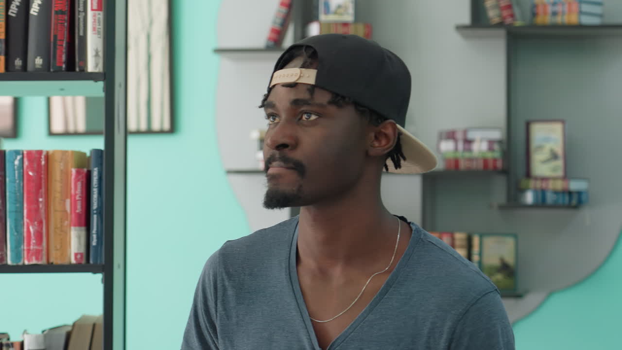 Contemplative young man in casual clothing and backward cap walking through library, surrounded by shelves of colorful books, looking down with thoughtful expression