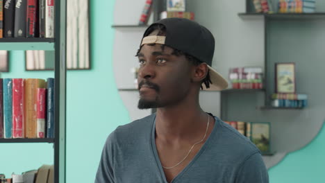 contemplative young man in casual clothing and backward cap walking through library, surrounded by shelves of colorful books, looking down with thoughtful expression