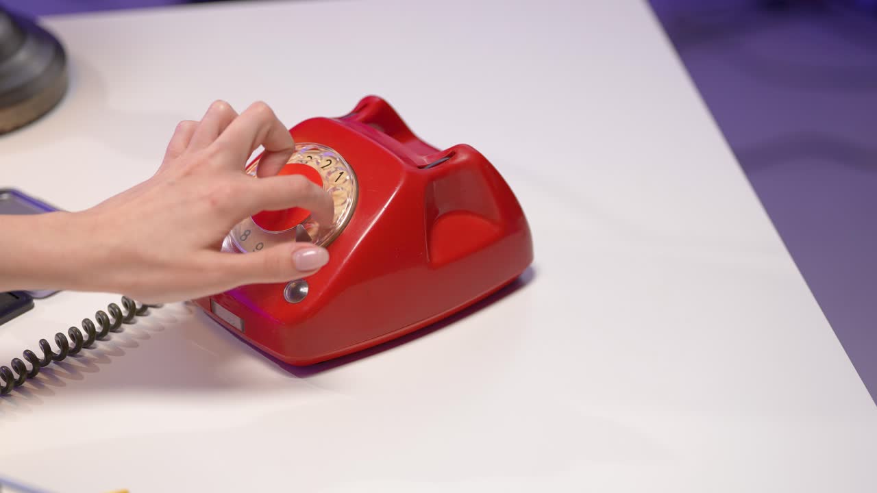 A close-up of a delicate hand reaching for the rotary dial of a bright red vintage phone. Placed on a clean white desk, this retro scene evokes classic communication in a stylish minimalist setting
