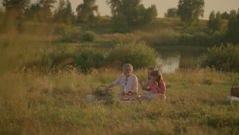 family seated on grassy field enjoying outdoor picnic, mother and middle-aged woman sharing moment with young girl reaching for glass of water, with background of greenery and small lake