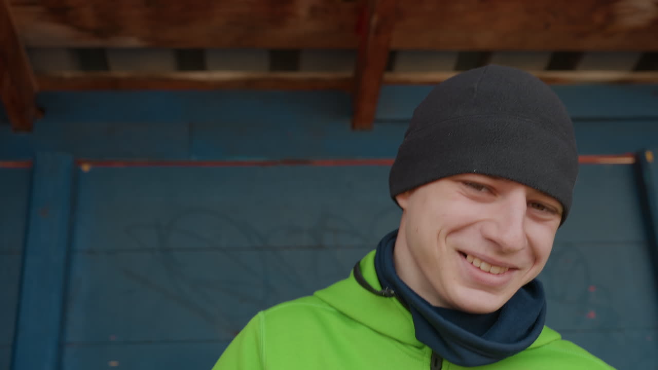 happy man under shelter smiling, man in beanie wearing bright jacket smiling happily, caucasian man enjoying genuine laughter beneath wooden shelter dressed in vivid green jacket and beanie