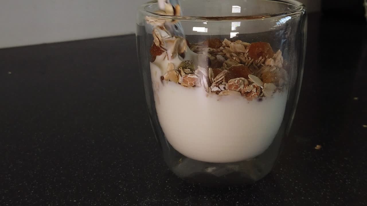 Yogurt mescolato con muesli e uvetta con un cucchiaio di metallo in una tazza di vetro.