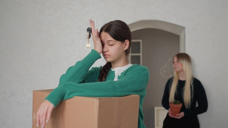 Young woman holding keys above box while contemplating exit, roommate in background with small plant, empty shelves and neutral walls, subtle shift from frustration to acceptance, quiet relief