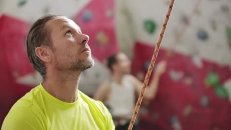 portrait of beautiful man rock climber belaying another climber with rope. indoors artificial climbing wall and equipment.