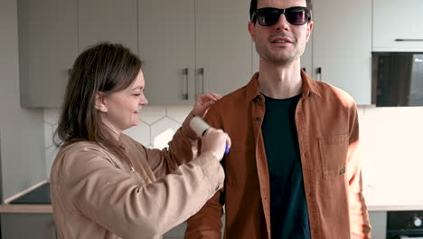 smiling woman helping a blind man to clean his clothes with lint roller while talking together at home