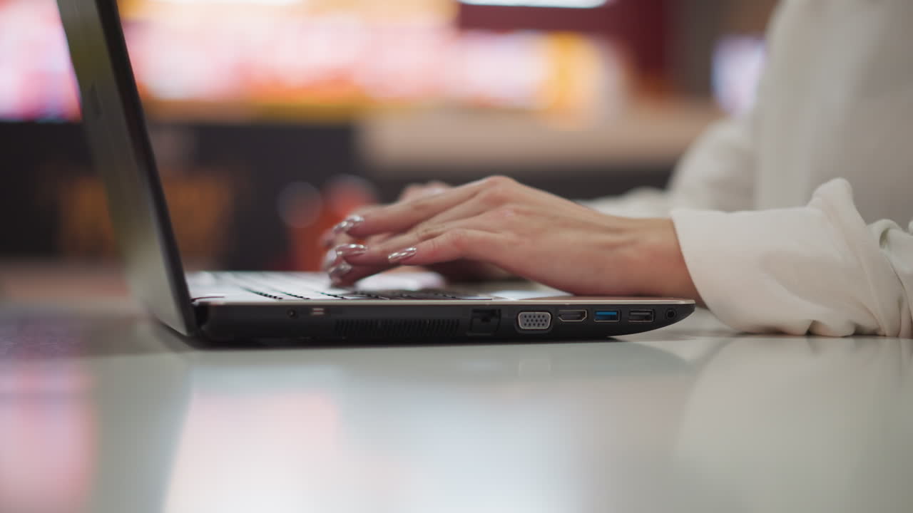 Elegant close-up of female hands typing on a laptop, showcasing painted nails and a professional work setting with blurred lights in the background