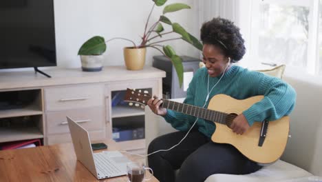 Feliz-Mujer-Afroamericana-Practicando-Guitarra,-Usando-Laptop-Y-Auriculares-En-Casa,-Cámara-Lenta
