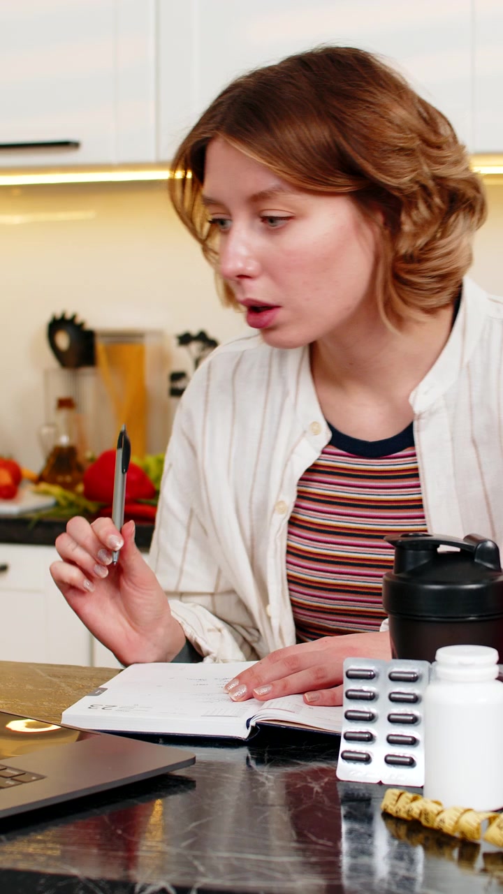 Giovane donna che prende appunti sulla nutrizione nella sua cucina di casa mentre pianifica i pasti con integratori sportivi.