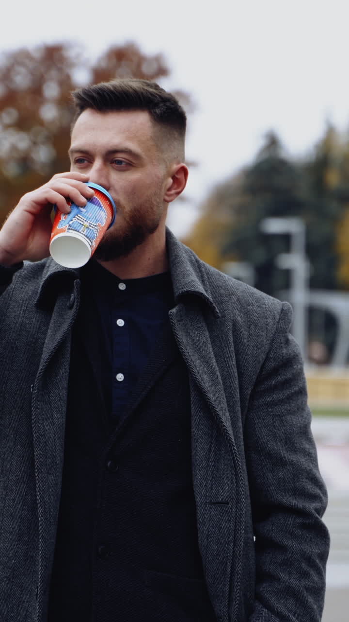 Young man drinking coffee in the street. Handsome bearded man in elegant warm coat walking in the city with a drink in plastic cup. Vertical video