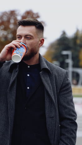 young man drinking coffee in the street. handsome bearded man in elegant warm coat walking in the city with a drink in plastic cup. vertical video