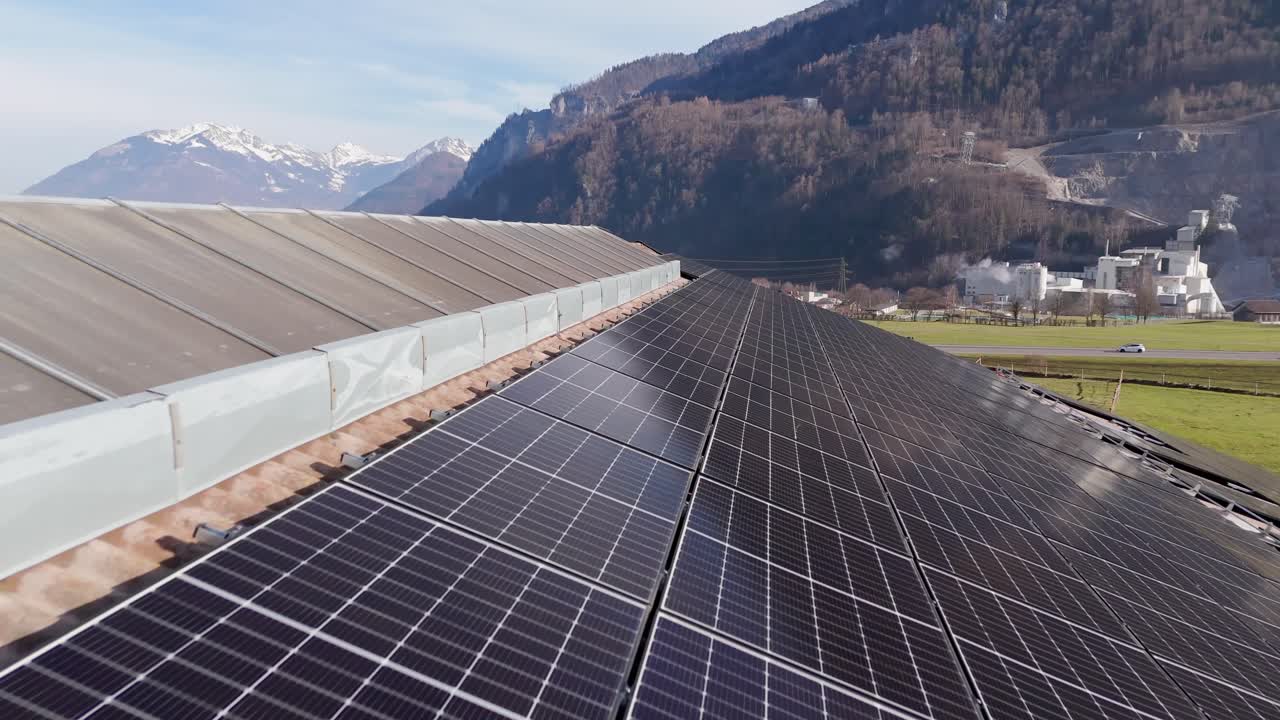 Aerial flight over solar panels installed on roof of barn on farmstead in Switzerland. Cars on highway during snowy winter mountains. Sunny day with snowy peaks of Alps. Wide shot.