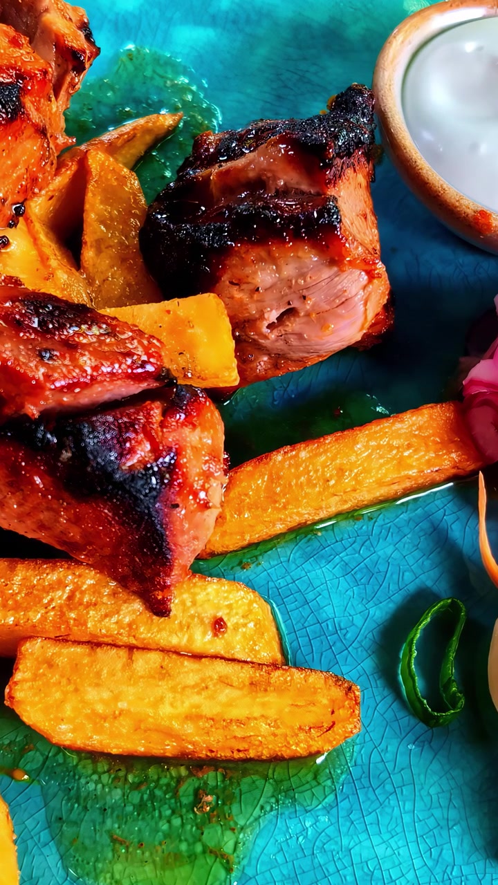 Succulenti pezzi di carne alla griglia abbinati a patate a spicchi dorate su un piatto blu brillante. Una ciotola di salsa e guarnizioni completano il piatto. Adatto per promozioni culinarie o contenuti di ricette