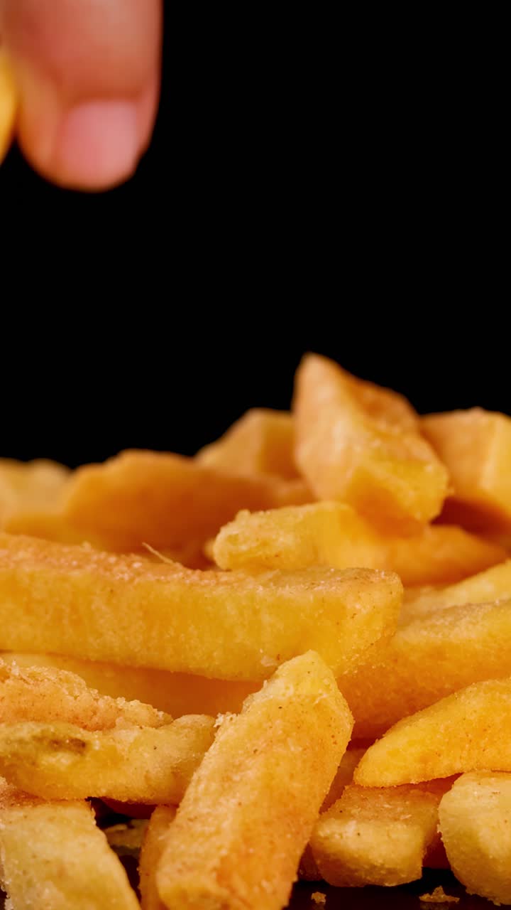Primer plano de una mano seleccionando patatas fritas doradas, iluminación dramática, fondo negro, movimiento suave de cámara.