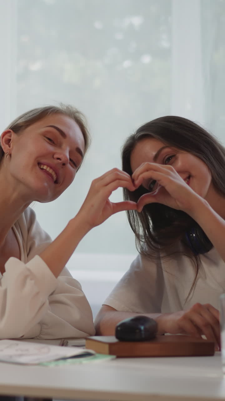 las mujeres lesbianas felices hacen forma de corazón con los dedos sonriendo y mirando a la cámara. la pareja lgbt muestra amor sentada en la mesa cerca de la computadora portátil en primer plano