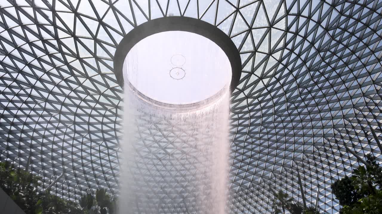 A dramatic indoor waterfall pours from a circular skylight in a glass-domed atrium, surrounded by tropical plants. Bright natural daylight, upward camera movement