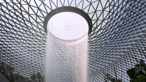 a dramatic indoor waterfall pours from a circular skylight in a glass-domed atrium, surrounded by tropical plants. bright natural daylight, upward camera movement