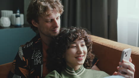 happy couple cudding on sofa at home, watching social media content on phone