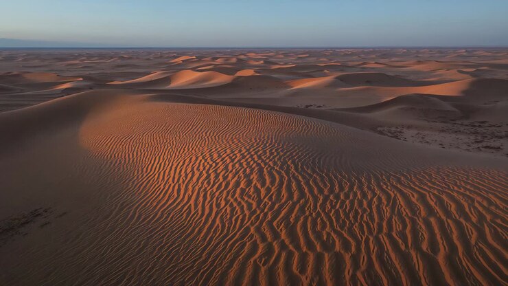 Ripresa aerea con drone in movimento che avanza sopra una duna desertica, mostrando creste e increspature illuminate dal sole