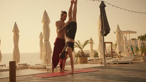 a brunette guy in red pants helps a girl in a black sports summer uniform stand on her hands head down on a red carpet on a sunny beach in the summer