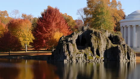 autumn landscape with rocky grotto, lake, and classical building