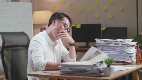tired asian man yawning and taking off his glasses before sleeping while working hard with documents at the office