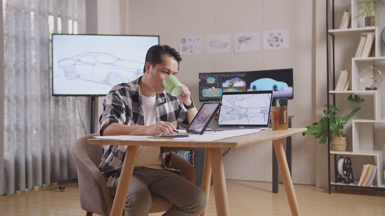 vista lateral de un hombre asiático bebiendo café mientras dibuja un nuevo concepto de diseño de automóvil en una tableta en el estudio con tv y computadoras muestra modelo de automóvil eléctrico 3d