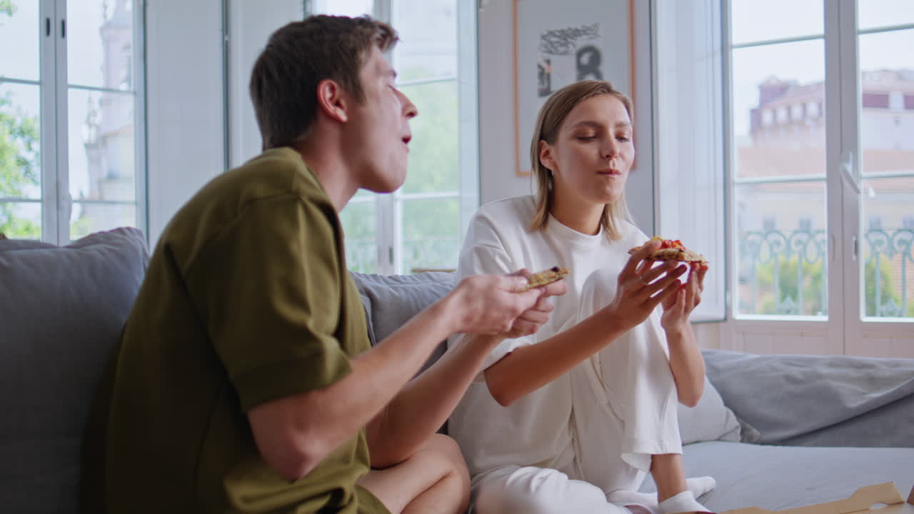 Hungry couple eating pizza sitting home sofa together. Happy people at lunch