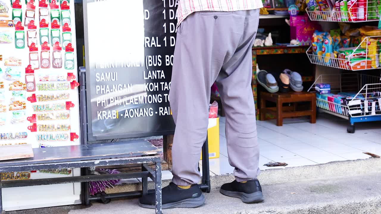 A vendor sweeps the sidewalk outside a colorful shop in Phuket, Thailand. Bright daylight illuminates the bustling street scene