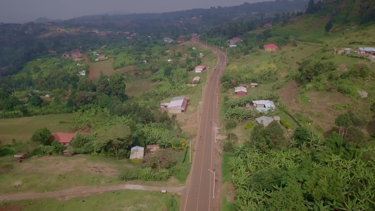 Drone shot of motorcycles riding on a paved road cutting through a lush, hilly rural landscape near Kapchorwa, Uganda, with scattered houses, vegetation, and forested hills in the background