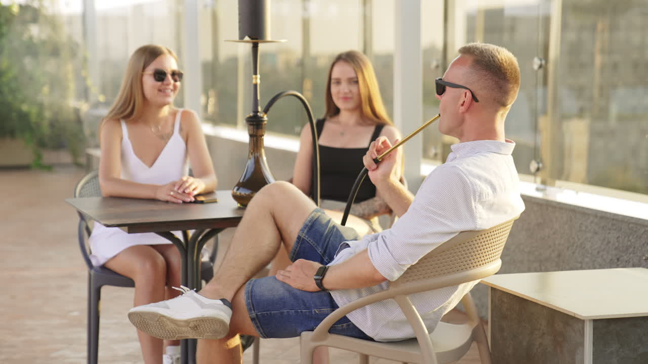 Company of happy relaxed people sitting outdoors at the table. Male smoking hookah and talking to female friends. Blurred backdrop.