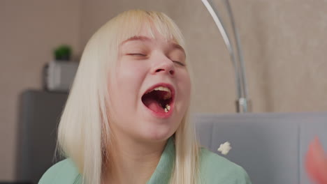 close up of blonde girl in green shirt smiling with satisfaction after catching popcorn in mouth during playful indoor moment, her joyful expression shows excitement as she enjoys the fun interaction