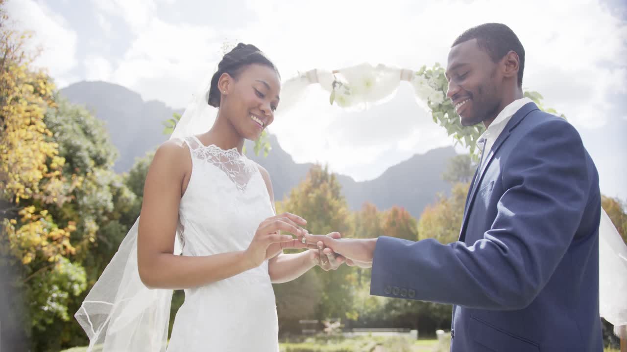 Happy african american bride putting ring on groom at wedding ceremony in sunny garden, slow motion