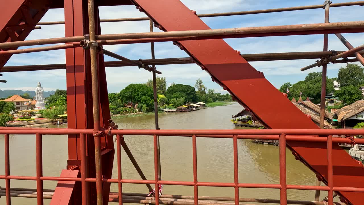 Camera travels across a red metal bridge under construction, revealing river, riverside buildings, and lush landscape in bright daylight, Kanchanaburi, Thailand