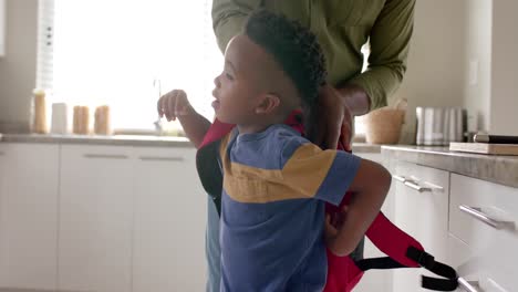 african american grandfather giving schoolbag to grandson in kitchen before school, slow motion