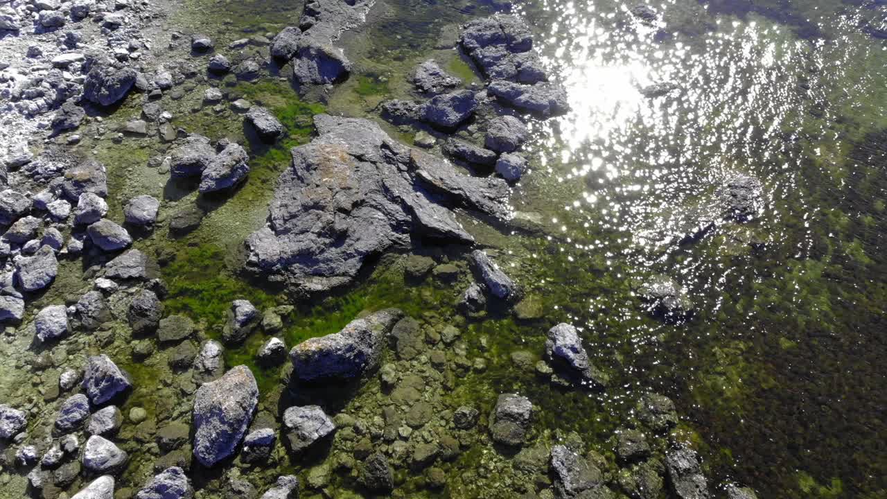 Aerial view over stones in shallow water, sun glittering on the water surface, on a warm, summer day, in Faro, Gotland, Sweden - low, tilt up,, drone shot