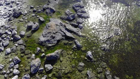 aerial view over stones in shallow water, sun glittering on the water surface, on a warm, summer day, in faro, gotland, sweden - low, tilt up,, drone shot