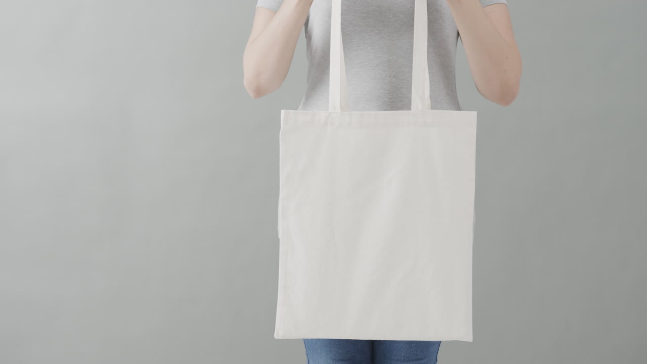 mujer caucásica con una camiseta blanca sosteniendo una bolsa blanca sobre un fondo gris, espacio de copia, cámara lenta