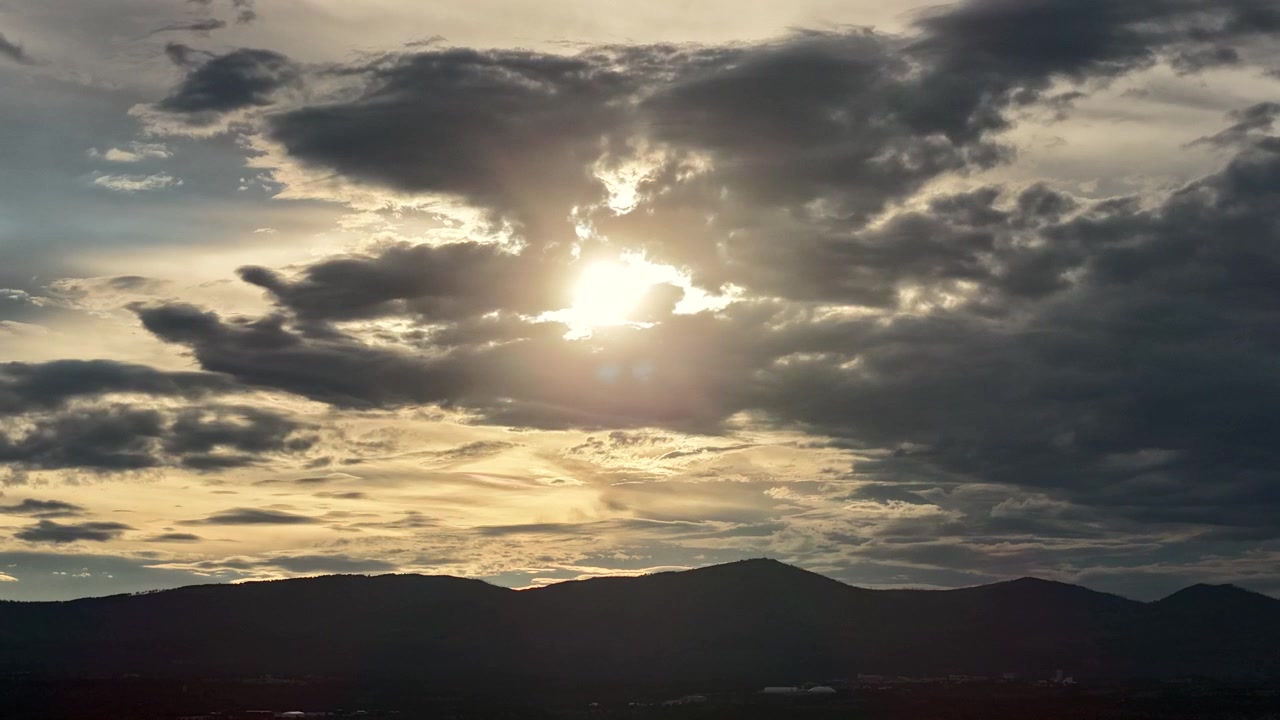 4K drone timelapse of the evening sun flickering behind thick, moody clouds. The overcast sky shifts over the dark silhouette of the Jemez Mountains behind Los Alamos, New Mexico.