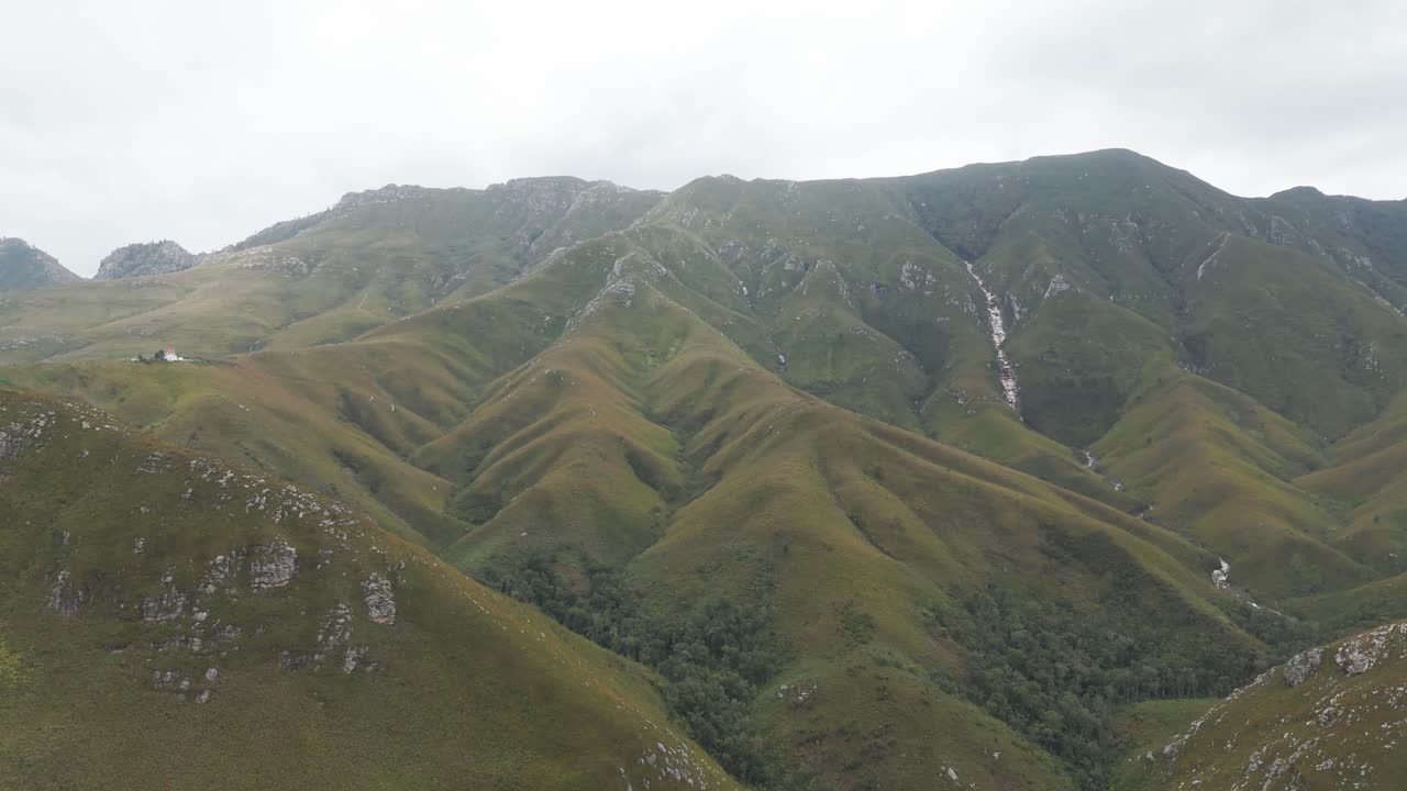 Aerial shot of green fynbos-covered mountains in Outeniqua Pass, Western Cape, South Africa. The drone moves down into the valley on a cloudy day, revealing layers of mountains in the misty backdrop