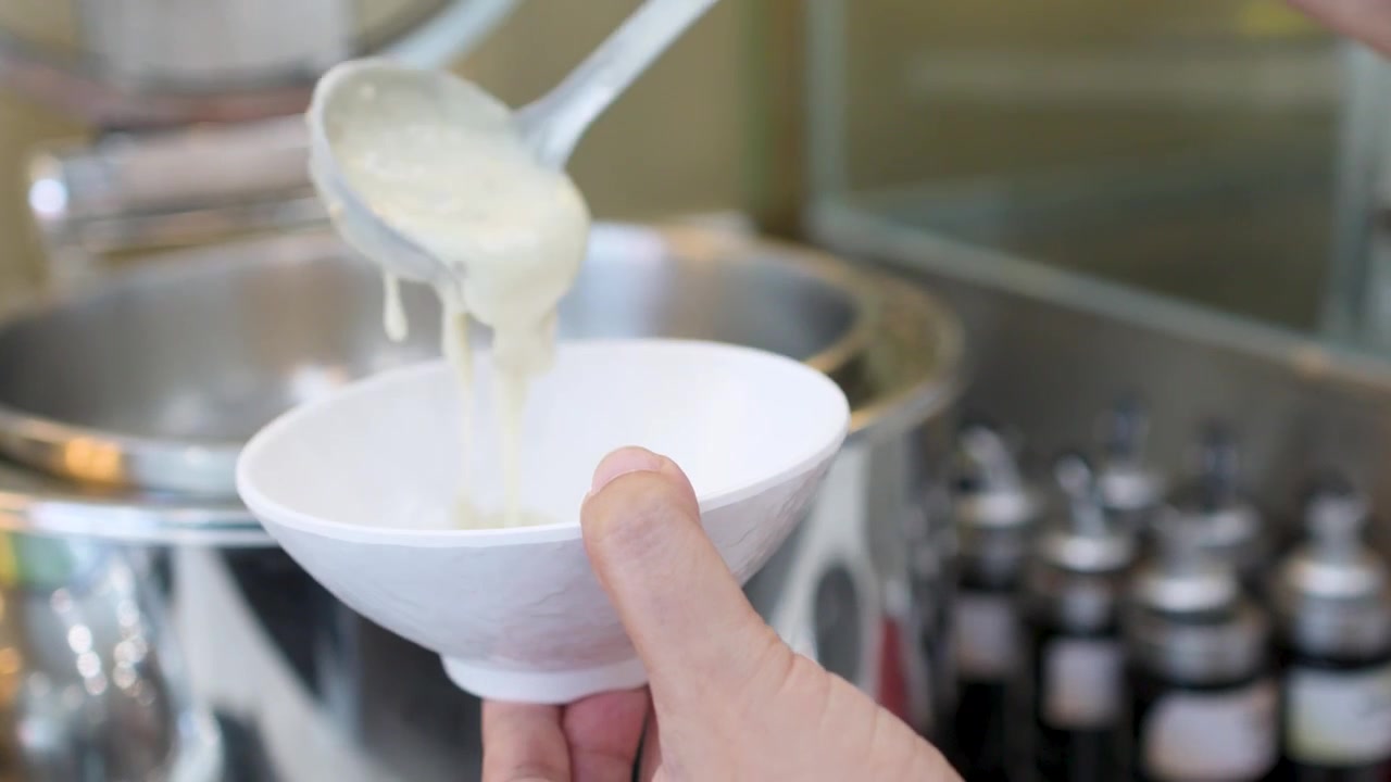 A hand ladles hot porridge from a metal pot into a white ceramic bowl
