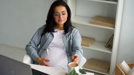 young happy pregnant business woman takes a break stroking her belly while having a hot drink