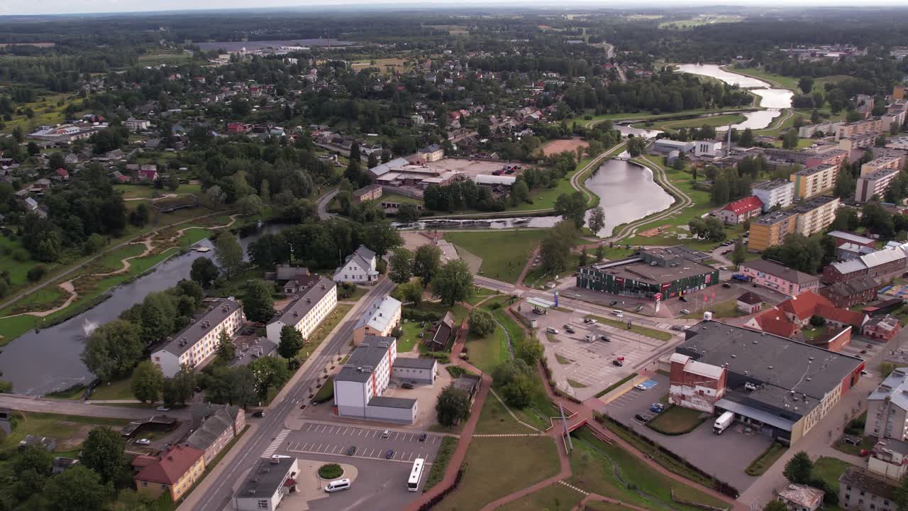 A drone captures an aerial view of the small town of Valka, showcasing its buildings, streets, and surrounding greenery. The peaceful townscape contrasts with the natural beauty of the area.