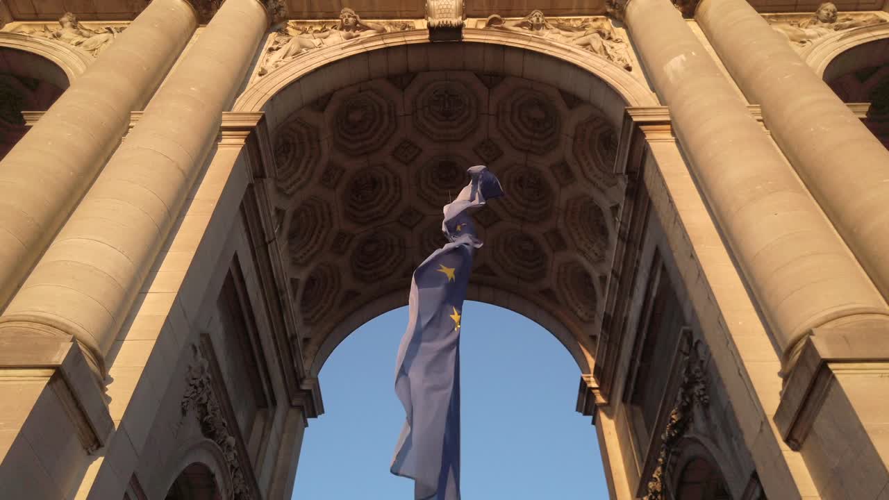 Dramatic low-angle shot of the European Union flag waving beneath the central arch of the Cinquantenaire monument in Brussels. The ornate coffered ceiling and stone columns frame the composition