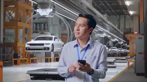 un trabajador profesional masculino asiático de pie con su teléfono inteligente en una fábrica de automóviles ev, una instalación industrial de alta tecnología, observa mirando a su alrededor antes de concentrarse con su teléfono móvil