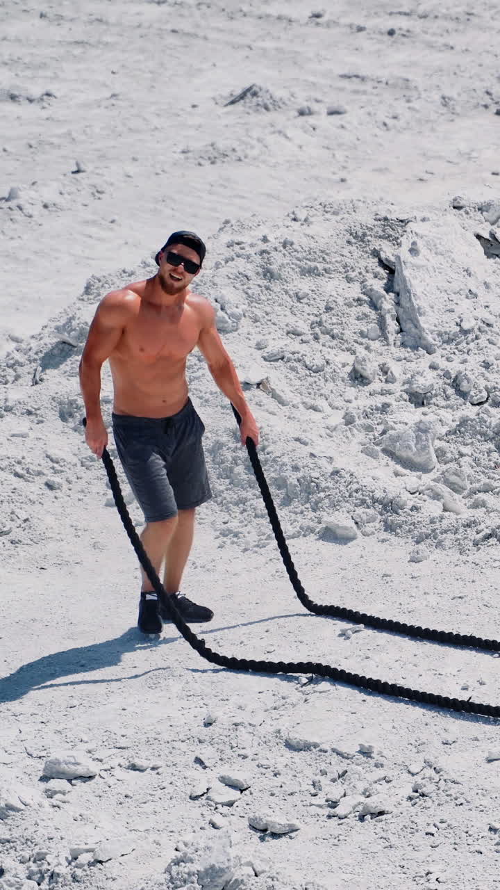 Happy sportsman finishing his workout with battle ropes outdoors. Topless athlete exercising with two ropes on the natural rocky background. Vertical video