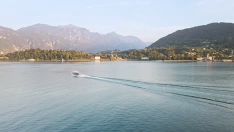 a stunning drone shot at lake como, moving forward as a boat heads towards bellagio. the serene lake and majestic mountains in the backdrop enhance the breathtaking scenery.