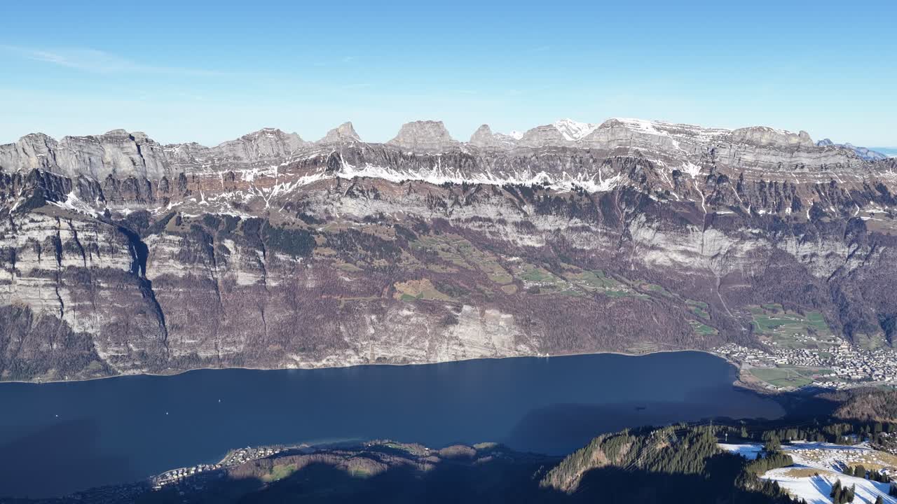 Bredt luftfoto af en alpinsø omgivet af dramatiske bjergklipper om vinteren, der viser et roligt naturlandskab under en klar, blå himmel