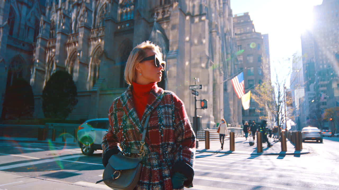 New York, USA, 25 ottobre 2025: Donna alla moda in cappotto a quadri vicino alla Cattedrale di San Patrizio. Donna bionda con occhiali da sole e dolcevita rosso in piedi su una strada di Manhattan con lo sfondo della cattedrale